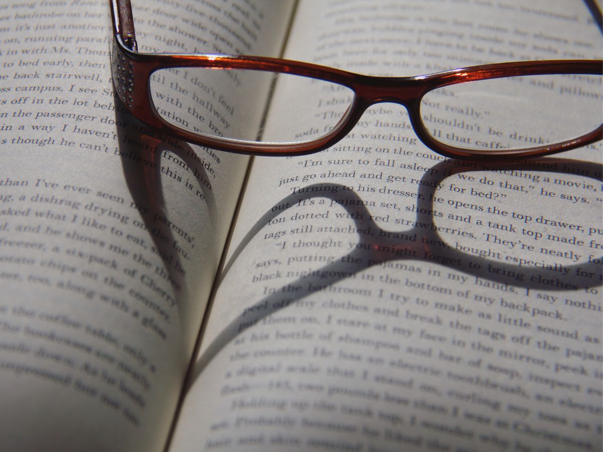 An open book, reading glasses resting on top, creating a heart-shaped shadow